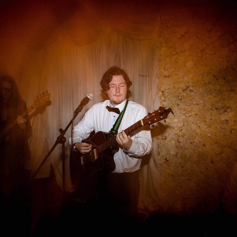 Hosh Harper playing guitar with a bow tie in a dimly lit room, To advertise his performance at Worcester's 31 The Cross Cafe, Friday 12th September - 6-8pm