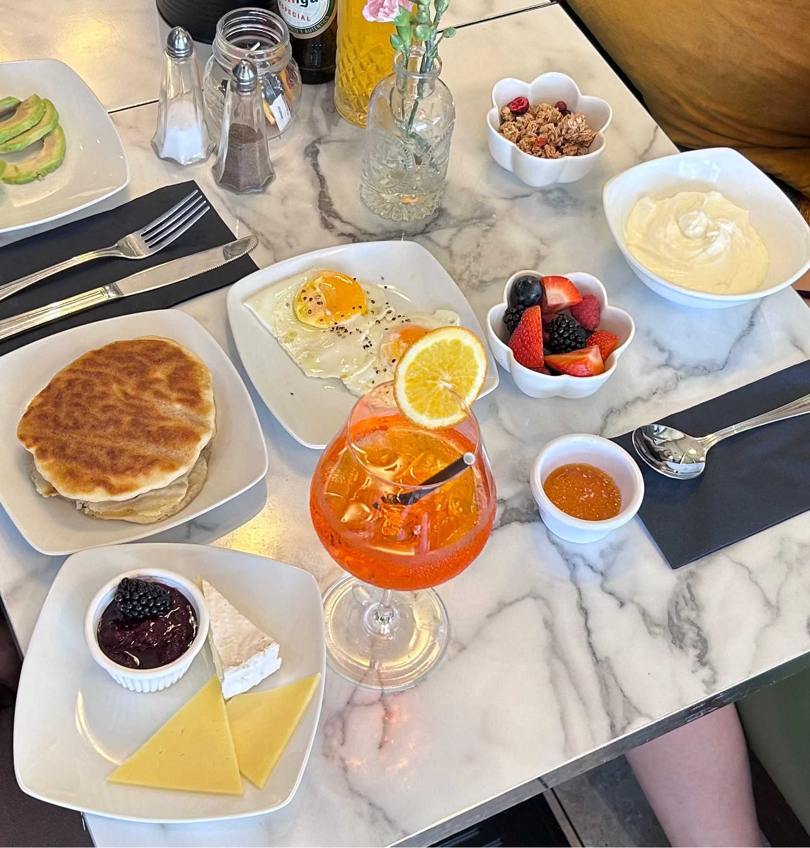 A breakfast spread on a white marble table, featuring pancakes, avocado slices, eggs with a sunny-side-up, fruit, yogurt, cheese, jam, and an orange cocktail in a glass. Parts of two people are visible around the table.